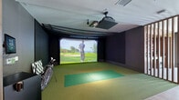 Located at 3421 N Freeway Blvd in Sacramento, CA, this thoughtfully designed amenity space combines a high-end golf simulator bay with a refined social lounge area. The space is divided into two distinct yet seamlessly connected zones, separated by a striking slatted wood partition that adds architectural character while maintaining an open, airy flow between areas.

The golf simulator bay features a large, high-definition projection screen, ceiling-mounted projector, and turf flooring in varying shades of green that recreate an authentic course feel. Dark, dramatic walls enclose the bay to enhance the immersive simulation experience, and golf bag storage is conveniently accessible nearby.

The adjacent lounge area is elegantly appointed with light hardwood-style flooring, tufted velvet sofas, tufted accent chairs, and layered patterned area rugs. Cluster pendant lights hang above the seating area, casting a warm ambiance. Decorative wall art, a mounted flat-screen television, and a potted olive tree contribute to the sophisticated atmosphere. Large windows allow natural light to filter in, offering views to the outdoors. Warm wood paneling and accents throughout the space unify the design, creating an inviting environment well-suited for both active recreation and relaxed social gatherings.
