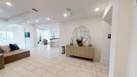 This single-story amenity space presents a cohesive coastal aesthetic, characterized by crisp white walls, light large-format tile flooring, and tray ceilings with recessed lighting throughout. The entry foyer sets an elegant tone with a gold geometric credenza topped by a round sunburst mirror and floral arrangement, providing a welcoming focal point upon arrival.

The lounge area features a striking tray ceiling with a vibrant turquoise accent, complemented by large windows that invite natural light and outdoor views. Leather club chairs and a sofa surround a glass-top coffee table atop a bold blue area rug, while oversized agate-inspired artwork adorns the walls.

A dedicated game room accommodates multiple round game tables, including a poker table, flanked by large-scale abstract paintings in deep navy tones. Expansive windows offer views of the surrounding community.

The fully equipped kitchen features white cabinetry, stainless steel appliances including double wall ovens and a refrigerator, a large island with pendant lighting, and a decorative blue mosaic tile backsplash with a wall-mounted television. An adjacent dining area provides additional seating.

A casual seating lounge displays a curated gallery wall of framed landscape artwork alongside plush sectional sofas. Restrooms and dual water fountains are conveniently located within the space.