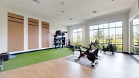 Located at 3175 E Corporate Center Pkwy in Kissimmee, Florida, this well-appointed fitness center offers a thoughtfully designed workout environment on a single floor. The space is defined by a clean, modern aesthetic, with crisp white walls, recessed ceiling lighting, and a combination of smooth hard flooring and vibrant artificial turf that delineates distinct workout zones.

Large, multi-pane windows line the exterior wall, flooding the room with natural light and providing open views of the surrounding greenery outside. The main workout area features a multi-functional training rack stocked with medicine balls, resistance bands, and assorted fitness accessories, alongside multiple stationary cycling bikes and cardio machines positioned for easy use. A wall-mounted flat-screen TV is ceiling-mounted for convenient viewing during workouts.

A dedicated turf zone provides space for agility drills, stretching, and functional movement exercises. Storage cubbies along one wall hold rolled yoga mats and step platforms, keeping the area organized and accessible. Striking hexagonal acoustic panels and athletic-themed wall decals add a dynamic, motivational character to the décor. Full-length mirrors along one wall enhance the sense of space and allow users to monitor their form. Glass-panel doors connect to adjacent areas, contributing to an open, airy feel throughout.