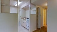 This single-floor residence offers a practical and functional layout designed to make the most of its available space. The kitchen is a standout area, featuring a built-in gas range, a sink with cabinetry below, and white lower cabinets that provide ample storage. A skylight positioned above the kitchen floods the area with natural light, creating a bright and airy atmosphere. Open shelving units adjacent to the kitchen offer additional storage and display space, adding both utility and character to the layout. The light-toned walls and neutral color palette throughout contribute to a clean and cohesive aesthetic. A hallway visible from the kitchen connects to the rest of the residence, hinting at additional rooms bathed in warm lighting. The flooring transitions from a light-toned surface in the kitchen area to a carpeted section further into the unit, adding a sense of warmth and comfort to the living areas. Ventilation is well considered, with visible air vents integrated into the walls. Overall, the space presents a straightforward and efficient design with thoughtful storage solutions and a welcoming ambiance enhanced by natural light.