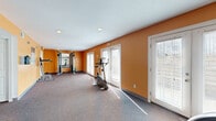 This fitness center is a well-organized, single-floor space designed to accommodate a variety of workout needs. The interior is immediately distinguished by its bold orange accent walls, which lend an energetic atmosphere throughout. The flooring is covered in durable, multi-toned carpet, providing a practical surface suited for exercise activity.

The main workout area is open and elongated, offering ample room to move between equipment. Cardio machines, including an elliptical and a stationary bike, are positioned to take advantage of natural light streaming in through several white-framed French doors fitted with horizontal blinds. These doors line the exterior wall, brightening the space and providing a visual connection to the outdoors.

At the far end of the room, a cable strength-training machine is anchored against the wall and accompanied by a wall-mounted mirror, allowing users to monitor their form during workouts. Additional equipment such as a rowing machine and free-standing benches round out the fitness offerings. Recessed ceiling lights supplement the natural light, ensuring the space remains well-lit throughout the day.

A secondary room, partially separated by a partial wall and doorway, provides additional utility space within the facility. An illuminated exit sign and safety panel are also visible, reflecting the space's compliance with safety standards. Overall, the fitness center presents a functional and motivating environment.