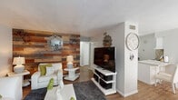 This single-floor space makes a strong design statement through a thoughtful blend of rustic and contemporary elements. The living area is the focal point, anchored by a striking reclaimed wood accent wall featuring multicolored planks in varying shades of gray, brown, and tan. Against this backdrop, crisp white seating — including a sofa and accent chair accented with green throw pillows — creates a bold contrast. A dark gray area rug grounds the arrangement, while a white media console and coffee table reinforce the modern aesthetic. Decorative mirrors and wall art complete the curated look.

The space transitions into a compact, well-organized kitchen featuring white cabinetry, a mosaic tile backsplash, and a peninsula with granite-style countertops that doubles as a breakfast bar with seating for two. Sheer curtains filter natural light into this area, maintaining a bright and airy feel throughout.

Wood-look flooring flows consistently through the main living areas, adding warmth and visual continuity. The bathroom offers a lively contrast with a bold, colorful bohemian-patterned shower curtain, a white vanity with a speckled countertop, a large mirror with Hollywood-style lighting, and warm-toned accent pieces that give the room a vibrant, personalized character.