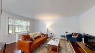 This single-floor residence offers a thoughtfully designed layout that balances comfort and functionality across its key living spaces. The living room serves as a welcoming centerpiece, featuring rich hardwood floors, light gray walls, and multiple windows that frame lush greenery outside, filling the space with natural light. The room accommodates a generous seating arrangement anchored by a patterned area rug, contributing to a warm, mid-century modern aesthetic.

The kitchen is well-equipped with white shaker-style cabinetry, classic subway tile backsplash, and granite countertops. A peninsula provides additional prep and seating space, while stainless steel appliances — including a gas range, dishwasher, refrigerator, and over-range microwave — add a modern edge. Dark tile flooring and pendant lighting complete the space. Multiple windows offer pleasant garden views.

Two bedrooms feature the same rich hardwood flooring found throughout the home. Both are bright and well-proportioned, with large windows overlooking mature trees and decorative radiator covers adding a classic architectural detail.

The bathroom is finished in clean white tile and includes a soaking tub with a handheld shower fixture, a natural light-filled window with outdoor views, and a crisp, functional layout throughout.