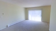 This single-floor space offers a clean, functional layout with a calm and understated aesthetic. The living area is a standout feature, characterized by its open feel and light-filled atmosphere. A large sliding glass door, dressed with vertical blinds, serves as the focal point of the room, inviting abundant natural light and providing outdoor access. The soft, neutral-toned carpeting underfoot adds warmth and comfort to the space, complementing the pale, cream-colored walls that contribute to an airy, open ambiance. Baseboard heating runs along the perimeter of the living area, ensuring efficient and consistent warmth throughout the colder months. The smooth, white ceiling and minimalist wall surfaces keep the space feeling fresh and versatile, providing a blank canvas well-suited for a variety of interior styles. Overall, the space presents a practical and welcoming environment, with its key features centered around natural light, comfortable flooring, and efficient heating — making it a straightforward yet comfortable living environment.