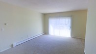 This single-floor space offers a clean, functional layout with a calm and understated aesthetic. The living area is a standout feature, characterized by its open feel and light-filled atmosphere. A large sliding glass door, dressed with vertical blinds, serves as the focal point of the room, inviting abundant natural light and providing outdoor access. The soft, neutral-toned carpeting underfoot adds warmth and comfort to the space, complementing the pale, cream-colored walls that contribute to an airy, open ambiance. Baseboard heating runs along the perimeter of the living area, ensuring efficient and consistent warmth throughout the colder months. The smooth, white ceiling and minimalist wall surfaces keep the space feeling fresh and versatile, providing a blank canvas well-suited for a variety of interior styles. Overall, the space presents a practical and welcoming environment, with its key features centered around natural light, comfortable flooring, and efficient heating — making it a straightforward yet comfortable living environment.