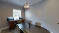 This single-floor business space is designed to support both collaborative meetings and individual work. At its heart sits a large, dark oval conference table surrounded by cushioned chairs, making it well-suited for team gatherings or professional meetings. A striking spherical chandelier with exposed Edison-style bulbs hangs above the table, adding a refined yet contemporary touch to the room's ambiance.

The space is finished with neutral gray walls accented by crisp crown molding, lending a clean and polished aesthetic throughout. A bright window fitted with white horizontal blinds allows natural light to filter into the room, creating a comfortable and inviting atmosphere.

Along one wall, a dedicated workstation area is equipped with desktop computers and additional seating, offering a practical setup for individual tasks or focused work sessions. The combination of meeting and computing resources makes this a versatile space suited for a variety of professional needs.

Recessed lighting complements the chandelier overhead, ensuring the room is well-lit at all times. The overall design balances functionality with a sophisticated style, making it an ideal environment for productivity and professional interaction.