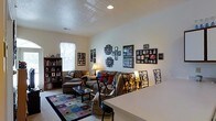 This single-floor space offers a comfortable and well-utilized layout centered around an inviting living area. The room features a textured ceiling with recessed lighting, providing a clean, well-lit environment. A large window along one wall allows natural light to brighten the space, complementing the warm, neutral wall tones throughout.

The living area is furnished with a plush sofa and loveseat arrangement, accented by colorful throw pillows that add vibrancy to the room. A glass-topped coffee table sits atop a patchwork-style area rug, adding both texture and visual interest to the floor space. The wall serves as a prominent display area, featuring an extensive and personalized gallery of framed photographs arranged in an eclectic, eye-catching collage.

A bookcase filled with books and decorative items lines one wall, adding a sense of character and function to the room. Wrought iron decorative panels provide an elegant visual divider between the living and dining areas. The dining space is partially visible, featuring ornate metal-backed chairs and a light-colored table, tying the overall aesthetic together with a blend of casual comfort and decorative detail.