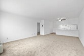 This single-floor, one-bedroom, one-bathroom unit offers a clean and functional layout across a comfortable amount of living space. The open-concept kitchen and living room form the heart of the unit, with the kitchen featuring white cabinetry, white countertops, a peninsula with additional counter space, a built-in cooktop, a white refrigerator, and a stainless sink beneath a window that lets in natural light. A ceiling fan with a light fixture overhead adds practical airflow to the combined living area, which is finished with neutral carpet throughout. Large windows in the living area provide outdoor views and fill the space with natural light. The bedroom is a well-proportioned, carpeted room with its own window offering natural light and outdoor views, and connects to a dedicated closet for storage. A hallway links the main living spaces to the private areas of the unit. The full bathroom is finished with wood-look flooring, a wide white vanity countertop with a large mirror, a bathtub/shower combination, and a window for natural ventilation. Throughout the unit, white walls and clean finishes create a bright, neutral palette well-suited for a variety of personal styles.
