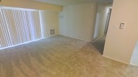 This single-floor space features an open living area finished with wall-to-wall neutral-toned carpeting that extends throughout, contributing to a quiet and comfortable atmosphere. The room is generously proportioned, providing ample space for a variety of furniture arrangements. A sliding glass door lined with vertical blinds runs along one wall, allowing natural light to fill the interior and offering potential access to an outdoor area. The walls are painted in a soft, warm neutral tone that complements the carpeting and creates a cohesive, understated aesthetic. A floor-level HVAC vent is built into the baseboard area beneath the window, indicating a standard forced-air climate control system. A wall-mounted fixture provides additional lighting options, while the smooth, clean wall surfaces remain free of architectural ornamentation, giving the space a straightforward and functional character. An adjoining area or hallway is visible toward the rear of the space, suggesting additional rooms or storage beyond the main living zone. Overall, the layout is practical and well-proportioned, with a clean, move-in-ready condition throughout.