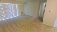 This single-floor space features an open living area finished with wall-to-wall neutral-toned carpeting that extends throughout, contributing to a quiet and comfortable atmosphere. The room is generously proportioned, providing ample space for a variety of furniture arrangements. A sliding glass door lined with vertical blinds runs along one wall, allowing natural light to fill the interior and offering potential access to an outdoor area. The walls are painted in a soft, warm neutral tone that complements the carpeting and creates a cohesive, understated aesthetic. A floor-level HVAC vent is built into the baseboard area beneath the window, indicating a standard forced-air climate control system. A wall-mounted fixture provides additional lighting options, while the smooth, clean wall surfaces remain free of architectural ornamentation, giving the space a straightforward and functional character. An adjoining area or hallway is visible toward the rear of the space, suggesting additional rooms or storage beyond the main living zone. Overall, the layout is practical and well-proportioned, with a clean, move-in-ready condition throughout.