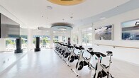 This well-appointed fitness studio offers a dedicated space for high-energy workouts in a clean, contemporary setting. The studio is designed with a crisp white color palette throughout, complemented by light herringbone-pattern flooring that lends a refined aesthetic to the athletic environment. A distinctive circular tray ceiling with teal blue accents serves as a striking architectural focal point, housing a modern pendant light fixture that adds an upscale touch to the space.

A full row of professional-grade Spinning bikes lines the center of the studio, providing ample capacity for group cycling classes. Freestanding heavy bags are positioned throughout the room, offering versatility for cross-training. A wall-mounted rack near the entrance stores additional fitness accessories, while a tiered stand holds a set of stability balls in varying sizes. Large flat-screen displays are mounted on the walls, and motivational artwork depicting outdoor adventure scenes adds visual energy to the space.

Floor-to-ceiling windows wrap around the exterior walls, flooding the studio with abundant natural light and providing open views of the surrounding outdoor area, including greenery and a pool courtyard beyond. A full-length mirror along one wall expands the sense of space and supports form-focused training. Glass sliding doors etched with "Fitness Studio" provide a transparent, modern entry point to this bright and inviting workout environment.