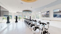 This well-appointed fitness studio offers a dedicated space for high-energy workouts in a clean, contemporary setting. The studio is designed with a crisp white color palette throughout, complemented by light herringbone-pattern flooring that lends a refined aesthetic to the athletic environment. A distinctive circular tray ceiling with teal blue accents serves as a striking architectural focal point, housing a modern pendant light fixture that adds an upscale touch to the space.

A full row of professional-grade Spinning bikes lines the center of the studio, providing ample capacity for group cycling classes. Freestanding heavy bags are positioned throughout the room, offering versatility for cross-training. A wall-mounted rack near the entrance stores additional fitness accessories, while a tiered stand holds a set of stability balls in varying sizes. Large flat-screen displays are mounted on the walls, and motivational artwork depicting outdoor adventure scenes adds visual energy to the space.

Floor-to-ceiling windows wrap around the exterior walls, flooding the studio with abundant natural light and providing open views of the surrounding outdoor area, including greenery and a pool courtyard beyond. A full-length mirror along one wall expands the sense of space and supports form-focused training. Glass sliding doors etched with "Fitness Studio" provide a transparent, modern entry point to this bright and inviting workout environment.