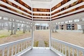 This classic outdoor gazebo is situated on the grounds of 456 S Main St in North Syracuse, New York. The structure features a traditional octagonal design finished in crisp white paint, with an open-air layout that allows for unobstructed panoramic views of the surrounding landscape. The gazebo's ceiling is composed of rich, warm-toned wood planks arranged in a radial pattern, providing a striking natural contrast to the white-painted framework. Elegant curved corner brackets and decorative spindle railings run along the perimeter, adding architectural character and charm to the structure. The wood-planked floor provides a sturdy and defined interior space. A walkway leads directly to the gazebo's entrance, connecting it to the broader grounds. The surrounding environment features a mix of mature deciduous trees, open grassy areas, and leaf-covered ground, indicative of a late-autumn or early-winter setting. Views from within the gazebo extend outward to the wooded hillside in one direction and to multi-story brick residential buildings in another. The gazebo serves as a functional and aesthetically pleasing outdoor gathering point within the property's communal green space.