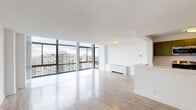 This single-floor residence is designed with a clean, modern aesthetic throughout, featuring a consistent palette of white walls and light wood-tone flooring that unifies the space. The expansive living area is a standout, defined by a sweeping wall of floor-to-ceiling windows fitted with dark metal frames, flooding the interior with natural light and offering elevated views of the surrounding cityscape and green treetops beyond.

The open-plan layout connects the living area to a peninsula-style kitchen, which features a galley configuration with olive-toned cabinetry, light countertops, stainless steel appliances including a gas range, microwave, and dishwasher, and a undermount sink. The contrast of the cabinetry against the white counters adds a contemporary touch.

The bedrooms are generously proportioned, each featuring ceiling fans, large windows with city views, and dedicated closet space — including both bifold and standard swing-door closets with built-in rods.

The bathroom includes a dark wood-toned vanity with a white countertop, an integrated mirror, white subway tile wainscoting, and a bathtub. Throughout, the space maintains a bright, minimalist character suited to modern living.