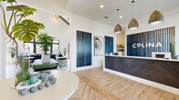 This expansive single-floor community clubhouse spans a generously sized footprint and is thoughtfully designed to serve as a central hub for residents. The entrance welcomes visitors with a sleek reception desk clad in a dark herringbone wood panel, pendant lighting, and bold blue slatted wall accents that set a contemporary coastal tone throughout. Light wood-look flooring flows seamlessly through the main areas, complemented by crisp white walls and abundant natural light.

The open lounge area features soaring vaulted ceilings with exposed wood beams, a dramatic sculptural pendant light, and a communal bar-height wood table positioned against a botanical-print accent wall with a built-in beverage station. Curated seating areas throughout the lounge incorporate curved upholstered chairs, natural wood coffee tables, and lush tropical plants, creating a warm and inviting atmosphere.

A dedicated leasing or welcome office offers a private consultation area with glass-paneled doors, wall-mounted display screen, and access to adjoining rooms. The well-appointed fitness center includes a full array of cardio machines, free weights, strength equipment, ceiling fans, mirrored walls, and a motivational typographic mural. A bright mail room with herringbone tile flooring, pendant lighting, and built-in mailbox banks rounds out the amenities. Large black-framed glass doors open directly to an outdoor pool area, filling the space with natural light and seamless indoor-outdoor connectivity.