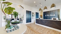 This expansive single-floor community clubhouse spans a generously sized footprint and is thoughtfully designed to serve as a central hub for residents. The entrance welcomes visitors with a sleek reception desk clad in a dark herringbone wood panel, pendant lighting, and bold blue slatted wall accents that set a contemporary coastal tone throughout. Light wood-look flooring flows seamlessly through the main areas, complemented by crisp white walls and abundant natural light.

The open lounge area features soaring vaulted ceilings with exposed wood beams, a dramatic sculptural pendant light, and a communal bar-height wood table positioned against a botanical-print accent wall with a built-in beverage station. Curated seating areas throughout the lounge incorporate curved upholstered chairs, natural wood coffee tables, and lush tropical plants, creating a warm and inviting atmosphere.

A dedicated leasing or welcome office offers a private consultation area with glass-paneled doors, wall-mounted display screen, and access to adjoining rooms. The well-appointed fitness center includes a full array of cardio machines, free weights, strength equipment, ceiling fans, mirrored walls, and a motivational typographic mural. A bright mail room with herringbone tile flooring, pendant lighting, and built-in mailbox banks rounds out the amenities. Large black-framed glass doors open directly to an outdoor pool area, filling the space with natural light and seamless indoor-outdoor connectivity.