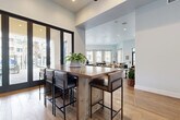 Located at 12406 Clark St in Moreno Valley, CA, this beautifully appointed single-story community clubhouse offers a generously sized, open-plan layout designed for both relaxation and socializing. The interior is defined by soaring ceilings, wide-plank hardwood flooring, and crisp white walls that create a bright, airy ambiance throughout.

The main gathering area flows seamlessly between distinct functional zones. A central lounge features a large sectional sofa, upholstered accent chairs, and a striking wood-paneled feature wall with a mounted flat-screen TV, lending a warm, contemporary character to the space. Adjacent to this is a game area anchored by a full-size pool table and additional game surfaces, ideal for casual entertainment.

A well-appointed bar and dining area showcases a substantial wood-top island with leather bar seating, set against a bold patterned tile accent wall and complemented by a full catering kitchen. A round dining table with upholstered chairs nearby adds a refined social setting.

Floor-to-ceiling black-framed glass doors and windows flood the interior with natural light and offer direct views and access to outdoor amenity areas. Recessed lighting throughout ensures the space is equally inviting after dark. The cohesive modern design, quality furnishings, and thoughtful layout make this clubhouse a standout community amenity.