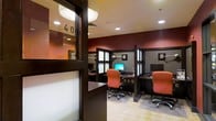 This business center is a single-floor facility designed to meet the productivity needs of its users. The space is thoughtfully laid out with multiple individual workstations, each separated by dark-stained wood privacy partitions that create a focused and semi-private working environment. Each station is equipped with a desktop computer and monitor, task lighting, and an ergonomic orange upholstered chair, ensuring comfort during extended work sessions. The rich, deep red accent walls add a bold and professional character to the space, complemented by the warm tones of the dark wood furnishings. Recessed lighting in the white ceiling provides consistent, even illumination throughout. Frosted glass panels integrated into the partition frames allow light to filter through while maintaining visual separation between stations. The flooring transitions between a light tile and carpeted mat areas beneath each workstation, adding both practicality and visual texture. A glass-paneled entry door with a numbered sign contributes to the organized, professional atmosphere of the room. Overall, the space strikes a balance between functionality and design, making it a well-appointed environment for business and personal computing tasks.