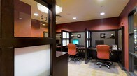 This business center is a single-floor facility designed to meet the productivity needs of its users. The space is thoughtfully laid out with multiple individual workstations, each separated by dark-stained wood privacy partitions that create a focused and semi-private working environment. Each station is equipped with a desktop computer and monitor, task lighting, and an ergonomic orange upholstered chair, ensuring comfort during extended work sessions. The rich, deep red accent walls add a bold and professional character to the space, complemented by the warm tones of the dark wood furnishings. Recessed lighting in the white ceiling provides consistent, even illumination throughout. Frosted glass panels integrated into the partition frames allow light to filter through while maintaining visual separation between stations. The flooring transitions between a light tile and carpeted mat areas beneath each workstation, adding both practicality and visual texture. A glass-paneled entry door with a numbered sign contributes to the organized, professional atmosphere of the room. Overall, the space strikes a balance between functionality and design, making it a well-appointed environment for business and personal computing tasks.