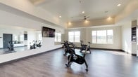 This single-floor fitness facility is thoughtfully designed to support a variety of workout routines, offering both a dedicated spin cycling studio and a separate yoga studio. The spin area is equipped with multiple Stages cycling bikes arranged for group classes, complemented by a wall-mounted flat-screen TV for guided workout content. Large windows along one wall flood the room with natural light and offer exterior views, while a ceiling fan maintains airflow throughout sessions. Full-length wall mirrors enhance the sense of space and allow participants to monitor their form during workouts. Recessed lighting throughout provides consistent, well-distributed illumination. The yoga studio is a clean, open space with ample room for movement and stretching, featuring the same durable hardwood-style flooring found throughout the facility. Crisp white walls, a tray ceiling with a ceiling fan, and a minimalist aesthetic create a calm, focused environment ideal for mindfulness and low-impact exercise. Dark-framed doors and bold dark baseboards provide a modern contrast to the light-toned walls. Together, the two spaces form a well-appointed fitness destination designed for both high-energy cycling sessions and restorative practice.