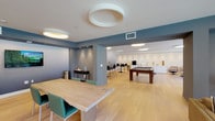 Located at 900 N La Brea Ave in Los Angeles, this contemporary amenity lounge delivers a polished, multi-functional environment designed for socializing, lounging, and recreation. The open-plan layout flows seamlessly across light hardwood flooring, unified by a clean modern aesthetic with cool blue-gray and white wall tones throughout.

The lounge area features plush leather sofas and upholstered armchairs arranged around a marble-topped coffee table, anchored by a bold navy area rug. Striking circular LED ceiling fixtures illuminate the space with warm, ambient light. Large floor-to-ceiling sliding glass doors flood the interior with natural light and open directly onto a furnished outdoor terrace with seating and greenery, offering views of the surrounding Los Angeles skyline.

A dedicated game and entertainment zone includes a full-size pool table and high-top bar seating arranged around a wall-mounted flat-screen TV, ideal for casual gatherings. A built-in kitchenette features sleek white cabinetry, a stainless steel double sink, and a white subway tile backsplash, providing convenient service support for events.

Additional seating vignettes include a live-edge communal table with teal accent chairs and a distinctive red upholstered booth, offering versatile settings for work or social use. The dollhouse view confirms the generous, well-proportioned footprint of this thoughtfully designed community amenity space.