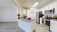 This single-floor residence is designed with a clean, modern aesthetic throughout, featuring light wood-look flooring and a crisp white color palette that creates a bright, cohesive atmosphere. The open-concept layout connects the kitchen and living area seamlessly, maximizing the sense of space and ease of movement.

The kitchen is a functional centerpiece, anchored by a peninsula island with a dark stone countertop that provides both prep space and casual seating. White shaker-style cabinetry lines the walls, offering ample storage, while stainless steel appliances — including a refrigerator, electric range, and over-the-range microwave — complete the setup. A flush-mounted ceiling light ensures the kitchen is well-illuminated. A dedicated laundry area houses a full-sized washer and dryer with overhead shelving for added convenience.

The bedroom benefits from multiple windows fitted with horizontal blinds, allowing natural light to filter through while maintaining privacy. A generously sized walk-in closet with dual hanging rods and shelving provides substantial storage capacity. The bathroom features a white vanity with stone countertop, a large mirror, and a shower-tub combination, maintaining the clean, neutral design carried throughout the space.
