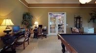 This single-floor space is designed as a versatile game and leisure room, combining entertainment and relaxation in a cohesive, well-appointed environment. The room is finished with warm golden-tan walls, crisp white crown molding, and neutral carpet, creating an inviting and polished atmosphere throughout.

A centerpiece pool table anchors the main entertainment area, complemented by a round glass-top bar-height table surrounded by leather-upholstered rattan chairs — ideal for casual dining or socializing. A wall-mounted flat-screen television is positioned above a dark wood credenza, which provides additional storage and display space. Botanical artwork adorns the walls, contributing to the room's relaxed, tropical-inspired aesthetic.

A cozy seating nook features a cushioned armchair framed by plantation shutters on multiple windows, allowing natural light to filter in while maintaining privacy. Lush indoor tropical plants are placed throughout, enhancing the organic, resort-like feel of the space.

A dedicated desk area with seating offers a functional workspace within the room. Elegant French doors with glass panes connect this space to adjacent areas, allowing light to flow through while providing a sense of openness and architectural charm.