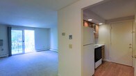 This single-floor space is thoughtfully laid out, offering a clear separation between the living area and kitchen while maintaining an open, connected feel. The living area is finished with wall-to-wall carpeting and features a baseboard heating unit along the exterior wall. A large window allows natural light to fill the room, creating a bright and airy atmosphere. Neutral, cream-toned walls throughout contribute to a clean and understated aesthetic.

The kitchen is positioned adjacent to the living area, separated by a partial wall that defines the space without fully enclosing it. It features warm-toned wood cabinetry, recessed lighting in the ceiling, and hardwood flooring that contrasts with the carpeted living space. Essential appliances are included, among them a white refrigerator and a range with an electric cooktop. A dishwasher is also integrated into the cabinetry. The kitchen's layout makes efficient use of its footprint, with upper and lower cabinetry providing ample storage. Overall, the space presents a functional and livable environment with a straightforward, practical design.