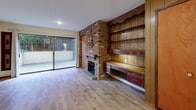 Located in Seattle's Greenwood neighborhood, this efficiently designed studio offers a thoughtful layout that maximizes every square foot. The combined kitchen and living area serves as the heart of the space, anchored by a striking floor-to-ceiling brick fireplace with a wood mantel — a standout architectural feature that adds warmth and character. Adjacent to the fireplace, a wall of rich wood-paneled built-in shelving and a built-in desk/credenza provide generous storage and display space. Light wood-tone flooring runs throughout, lending a clean, cohesive look. A sliding glass door floods the living area with natural light and opens directly onto a private patio, offering a seamless indoor-outdoor connection with views of surrounding greenery. The kitchen features warm wood cabinetry extending to the ceiling, a peninsula with a built-in cooktop and white countertops, a full-size refrigerator, and a sink — making it a functional and well-appointed cooking space. Two closets offer ample storage throughout the unit. The bathroom is clean and straightforward, with a tiled shower, vanity, and a grab bar. Baseboard heating ensures year-round comfort in this well-maintained, character-filled Seattle studio.