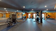 This single-floor fitness center is designed to support a full range of workout activities, offering a thoughtfully organized layout across multiple interconnected zones. The space is characterized by bold orange accent walls, dark ceilings with recessed lighting, and durable rubber flooring in the main training areas — with colorful patterned sections adding visual energy to the space.

The primary strength and cardio area is generously proportioned, housing cable machines, Precor-branded equipment, ellipticals, stationary bikes, and other cardio machines positioned near windows that allow natural light to filter in. Floor-to-ceiling mirrors line portions of the walls, enhancing the sense of openness and allowing users to monitor their form.

A separate group exercise and studio space features smooth hardwood-style flooring, large wall mirrors, and glass partitions, making it well-suited for yoga, cycling classes, or group fitness sessions. Spin bikes are positioned along one side, and foam rollers and step platforms are stored neatly along the mirrored wall.

A transitional area connects the zones and serves as a functional training hub, with wall-mounted stability ball racks stocked with exercise balls in various sizes, resistance bands, balance discs, and additional accessories. Accordion-style room dividers offer flexible configuration of the studio space. The overall ambiance is vibrant, energetic, and purpose-built for fitness.