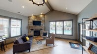 This single-floor clubhouse is thoughtfully designed to serve as a comfortable and functional gathering space. The main lounge area immediately draws attention with its soaring vaulted ceiling accented by a rustic exposed wood beam and an eye-catching wagon wheel chandelier overhead. The focal point of the room is a floor-to-ceiling natural stone fireplace with a reclaimed wood mantel and a wall-mounted flat-screen TV, creating a warm and inviting atmosphere. Plush seating, including a sectional sofa and accent chairs, is arranged around a round coffee table atop a woven area rug, encouraging conversation and relaxation. Large windows look out toward what appears to be a pool area, filling the space with natural light. Light wood-look flooring runs throughout, complementing the gray walls and overall neutral palette. Adjacent to the lounge, a well-appointed kitchenette features rich warm-toned wood cabinetry, dark granite countertops, a patterned tile backsplash, a stainless steel refrigerator, and a central island with bar seating. A casual high-top dining area is positioned nearby. The space also includes package lockers and a workstation area, adding practical convenience to the overall amenity-rich environment.