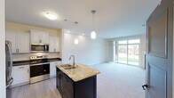 This single-floor residence offers a thoughtfully designed open-concept layout that seamlessly connects the kitchen and living area. The kitchen is a standout feature, showcasing white raised-panel cabinetry with dark hardware, dark countertops along the perimeter, and a large central island topped with granite countertops and an integrated double-basin sink. Elegant pendant lighting hangs above the island, while a full suite of stainless steel appliances — including a French door refrigerator, electric range, and built-in microwave — provides a complete and functional cooking environment. Light wood-look flooring runs throughout the kitchen, transitioning into plush carpeting in the living areas. The spacious living room is anchored by a fireplace with a tile surround and mantle, and a large sliding glass door with transom windows floods the space with natural light while offering views of the surrounding greenery. The bedroom is generously sized with soft carpeting, natural light from a double window, and direct access to a full bathroom. The bathroom is well-appointed with a dark wood vanity, white countertop, a large framed mirror, and a shower-tub combination. Throughout the residence, a neutral color palette and clean finishes create a bright, welcoming ambiance.