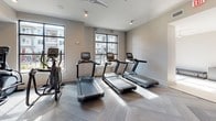 Located at 5520 Keltonwood Rd in Charlotte, NC, this single-floor fitness center is thoughtfully designed to accommodate a wide range of workout routines and fitness goals. The space is organized into several interconnected zones, each dedicated to a specific type of exercise, allowing users to move seamlessly between training modalities.

The main strength and functional training area features a multi-functional cable machine, a full rack of dumbbells, a weight bench, and a stair climber, all positioned beneath a wall-mounted television for entertainment during workouts. An adjacent section is devoted to functional fitness accessories, including a well-stocked rack of medicine balls, kettlebells, resistance bands, and a heavy punching bag, catering to dynamic and high-intensity training styles.

A dedicated cardio zone houses multiple treadmills and rowing machines, bathed in natural light streaming through large, floor-to-ceiling windows that frame outdoor views. A separate spin area accommodates stationary cycling bikes, offering an additional space for low-impact cardiovascular training.

Throughout the facility, patterned herringbone carpet tiles provide a clean, modern aesthetic, while recessed lighting and ceiling fans maintain a comfortable environment. The open, airy layout and neutral color palette contribute to a bright, welcoming atmosphere well-suited for daily exercise.