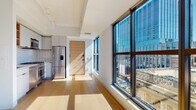 This contemporary single-floor unit offers a thoughtfully designed open layout bathed in abundant natural light, courtesy of expansive floor-to-ceiling windows that frame striking urban skyline views. Light hardwood flooring runs throughout, lending warmth and continuity to the space.

The open living area flows seamlessly from the entry, anchored by large picture windows that allow sunlight to stream in and provide unobstructed views of surrounding city architecture. A structural column adds a subtle architectural detail to the living zone.

The kitchen is well-appointed with a full suite of stainless steel appliances, including a Fisher & Paykel gas range, built-in microwave, dishwasher, and a French door refrigerator. White cabinetry is complemented by open warm wood shelving and a distinctive hexagonal white tile backsplash, creating a clean yet inviting aesthetic. Ample counter space and storage make the kitchen both functional and visually refined.

The bedroom features its own generous window wall overlooking city rooftops, maintaining the unit's bright, airy character. The bathroom is finished with contrasting dark hexagonal mosaic floor tiles, white penny-tile shower surround, a soaking tub, and a floating wood-accented vanity with integrated storage, delivering a polished, modern aesthetic throughout.