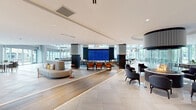 Located at 4 Vermella Way in Union, New Jersey, this striking amenity lounge is a masterfully designed communal space that blends contemporary architecture with upscale interior design. The single-floor layout is exceptionally spacious, characterized by soaring double-height ceilings in key areas that amplify the sense of openness and grandeur throughout.

Large-format light stone tile flooring flows seamlessly across the entire space, accented by integrated linear LED lighting that guides movement between zones. Floor-to-ceiling windows and glass doors line the perimeter, flooding the interior with abundant natural light and offering expansive views of the surrounding landscaped grounds and adjacent buildings.

The lounge is organized into several distinct functional areas. A central gathering zone features a large-scale video display wall flanked by dark architectural columns, surrounded by a mix of curved sectional sofas, upholstered lounge chairs, and dining-height seating in warm orange and charcoal tones. A dedicated recreation area includes foosball, billiards, and ping pong tables beneath dramatic pendant light fixtures. A quieter lounge section offers blue accent chairs and intimate seating arrangements, while a separate area showcases contemporary artwork and a fireplace-style feature wall. The space also includes a bar-height counter seating area and an elegant dining table positioned near glass entry doors with direct outdoor access, completing this versatile, amenity-rich environment.