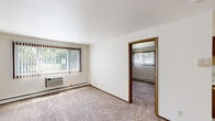 This single-floor unit offers a straightforward and functional layout spread across several well-defined rooms. The living area is generously sized, featuring plush wall-to-wall carpeting, crisp white walls, and warm wood trim that frames the doorways and baseboards throughout. Large windows fitted with vertical blinds allow natural light to fill the space while offering views of surrounding greenery. A wall-mounted HVAC unit and baseboard heating provide year-round climate control. An open doorway connects the living area to an additional carpeted room, ideal for use as a bedroom or flex space, which includes two closets with wood-trimmed doors. The kitchen is equipped with essential appliances including a white electric range, refrigerator, and a single-basin sink. Rich wood cabinetry lines the upper walls for ample storage, while tile flooring and a ceiling fan with a light fixture lend the space a practical and comfortable feel. Baseboard heating keeps the kitchen warm. The bathroom is clean and well-appointed, featuring a full tub-shower combination with tiled walls, a single vanity with an integrated sink, a framed mirror, overhead cabinetry with glass-front doors for additional storage, and tile flooring that ties the room together neatly.