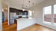 This thoughtfully designed single-floor space showcases a cohesive modern aesthetic highlighted by warm hardwood flooring and a bright, neutral palette. The open-concept kitchen serves as the centerpiece, featuring dark cabinetry, stainless steel appliances, a hexagonal tile backsplash, and a generous white quartz peninsula with a matte black sink fixture. Geometric pendant lights hang at varying heights above the peninsula, adding visual interest while illuminating the space. A track lighting system further enhances the kitchen's functionality and style. Adjacent to the kitchen, additional cabinetry and counter space with a window overlooking neighboring rooftops provide ample storage and natural light. The bedrooms are bright and airy, each benefiting from large windows that frame urban skyline views, filling the rooms with abundant natural light. Two well-appointed bathrooms feature slate-blue vanities with white countertops, classic basketweave tile flooring, matte black hardware and fixtures, and large mirrors. One bathroom includes a full soaking tub, while the other offers a refined half-bath layout. The overall design balances contemporary style with practical functionality, creating an inviting and cohesive living environment throughout.