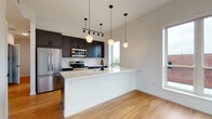 This thoughtfully designed single-floor space showcases a cohesive modern aesthetic highlighted by warm hardwood flooring and a bright, neutral palette. The open-concept kitchen serves as the centerpiece, featuring dark cabinetry, stainless steel appliances, a hexagonal tile backsplash, and a generous white quartz peninsula with a matte black sink fixture. Geometric pendant lights hang at varying heights above the peninsula, adding visual interest while illuminating the space. A track lighting system further enhances the kitchen's functionality and style. Adjacent to the kitchen, additional cabinetry and counter space with a window overlooking neighboring rooftops provide ample storage and natural light. The bedrooms are bright and airy, each benefiting from large windows that frame urban skyline views, filling the rooms with abundant natural light. Two well-appointed bathrooms feature slate-blue vanities with white countertops, classic basketweave tile flooring, matte black hardware and fixtures, and large mirrors. One bathroom includes a full soaking tub, while the other offers a refined half-bath layout. The overall design balances contemporary style with practical functionality, creating an inviting and cohesive living environment throughout.