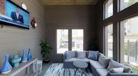 This thoughtfully designed single-floor space exudes a relaxed, coastal-inspired ambiance. The interior is clad in horizontal shiplap-style paneling in a deep charcoal tone, complemented by a matching wood-paneled ceiling that adds warmth and texture throughout. Large windows along the walls flood the space with abundant natural light while offering views of an outdoor terrace beyond, creating a seamless connection between interior and exterior. The flooring features a vibrant geometric area rug in shades of blue, green, and white, anchoring the central seating arrangement. A generous L-shaped sectional sofa with light grey upholstery and blue patterned accent pillows serves as the focal point, styled around a round metal side table for a polished yet casual feel. Lush potted tropical plants are positioned in the corners, introducing natural greenery that softens the moodier tones of the walls. A decorative wall sconce adds a rustic accent, enhancing the layered, curated aesthetic of the room. The overall atmosphere is cozy yet sophisticated, making it an inviting retreat well-suited for lounging or casual gatherings.