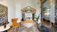 The Innova Clubhouse in Novi, Michigan is a thoughtfully designed, single-floor amenity facility offering a comprehensive range of community spaces. The welcoming entrance lobby features a striking combination of patterned carpet and warm hardwood-toned flooring, a dramatic green marble fireplace surround, coffered ceilings with elegant pendant lighting, and a bold leather lounge chair that sets a sophisticated tone. Adjacent lounge areas provide comfortable seating arrangements with floor-to-ceiling windows that invite abundant natural light and offer views of the outdoor pool and surrounding community. A well-appointed kitchen and entertaining area includes a green marble island, modern cabinetry, and full appliances, ideal for community gatherings. A dedicated media lounge features plush blue seating and a wall-mounted display screen. The expansive fitness center is equipped with treadmills, ellipticals, cycling machines, free weights, and cable systems, all beneath vibrant teal and orange accent ceilings with large windows overlooking the outdoors. A game room with a ping pong table opens directly to the pool area through oversized glass doors. A secure package room with smart lockers rounds out the facility's practical amenities.