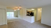 This single-floor space features an open-concept layout that connects the living area and kitchen, creating a cohesive and functional floor plan. The living area is finished with neutral wall-to-wall carpeting, contributing to a comfortable, understated atmosphere. A ceiling fan with an integrated light fixture is centrally mounted, providing both air circulation and ambient lighting. A full-length mirrored wall panel along one side of the living space adds a sense of depth and reflects natural light from a sliding glass door nearby, which offers access to an outdoor area and brings in additional daylight. The kitchen is positioned at the rear of the open space and is defined by a raised peninsula counter with a dark countertop, ideal for casual dining or food preparation. Medium-toned wood cabinetry lines the upper walls, and the kitchen is equipped with black appliances, including a refrigerator, microwave, and range. A flush-mount ceiling light provides dedicated illumination over the kitchen area. The overall color palette throughout is light and neutral, with white walls enhancing the bright, airy feel of the space.