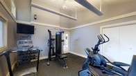 This single-floor fitness room offers a practical and functional workout space designed to accommodate a variety of exercise routines. The room features light grey walls accented by white crown moulding and baseboards, lending a clean and polished appearance throughout. Underfoot, durable dark carpet provides a stable, cushioned surface suitable for both machine-based and free-weight training.

The space is equipped with a multi-function cable resistance machine positioned centrally, offering users a wide range of strength training exercises. A dumbbell rack lines one wall, providing organized access to free weights, while a wall-mounted television adds entertainment options during workouts. On the opposite side, a BH elliptical trainer and additional cardio equipment round out the fitness offerings.

Overhead, exposed cylindrical ductwork runs along the ceiling alongside a ceiling fan, maintaining airflow and contributing to the room's utilitarian aesthetic. A glass-paneled door allows natural light to filter in, while recessed and overhead lighting ensures the space remains well-lit. The overall ambiance is straightforward and purposeful, prioritizing functionality and ease of use.