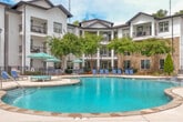 This expansive outdoor pool area offers a resort-style retreat designed for relaxation and leisure. At its center is a large, freeform pool with clear blue water, flanked by a wide concrete sun deck that accommodates numerous blue lounge chairs and shade umbrellas, providing ample space for sunbathing and outdoor gatherings. The pool's organic shape includes gentle curves and integrated entry points, with clearly marked depth indicators along the edges. Surrounding the pool, carefully maintained landscaping features ornamental evergreen trees, crepe myrtles, ornamental grasses, and mulched planting beds that add natural beauty and privacy to the space. Stone-clad architectural columns and structures frame the main entrance, lending a refined, upscale character to the area. Curved pathways guide residents through the landscaped grounds, connecting the pool deck to the surrounding two-story buildings, which feature balconies overlooking the courtyard. A dedicated outbuilding structure at the rear of the pool area provides additional amenity support. The entire space is enclosed and bordered by a lush tree line, creating a sense of seclusion while maintaining an open, airy atmosphere under the open sky.