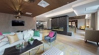 This sophisticated single-floor space is designed to impress, combining bold architectural elements with carefully curated furnishings to create a dynamic and inviting environment. The most striking feature is the richly textured, bronze-toned ceiling that spans the space, punctuated by recessed lighting and dramatic pendant fixtures that cast a warm, ambient glow throughout.

The layout is divided into distinct yet flowing zones. A generous lounge area anchors one end of the space, furnished with plush white sofas, vibrant yellow wingback chairs, and a mid-century-inspired charcoal accent chair, all arranged atop a patterned gold and yellow area rug. Large floor-to-ceiling windows and glass doors flood this area with natural light and offer views of the surrounding streetscape.

A sculptural latticed fireplace surround serves as a dramatic focal point, separating the lounge from the adjacent bar and social area. The bar features a sleek metallic island with a dark stone countertop, surrounded by teal and navy upholstered bar stools. Reclaimed wood accent walls display curated artwork, adding warmth and texture. A built-in media wall with a flat-screen TV and a discreet back kitchen complete the space, making it well-suited for both casual gatherings and private events.