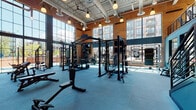 This two-floor fitness facility combines industrial architecture with a comprehensive suite of workout spaces. Exposed brick walls, soaring ceilings with visible ductwork, and floor-to-ceiling windows define the main weight training floor, flooding the space with natural light and offering views of the surrounding urban streetscape. The strength training area features multiple power racks, barbells, plate-loaded stations, and a full dumbbell rack arranged across resilient blue rubber flooring, all framed by bold athletic murals that energize the environment.

A dedicated cardio zone houses rows of treadmills, elliptical trainers, and stair climbers positioned along large windows, maximizing natural light during workouts. Warm hardwood flooring transitions the space from high-intensity zones into more functional training areas, including a room equipped with rowing machines and a dramatic black-and-white fitness mural. A versatile studio space features hardwood floors, large wall mirrors, spin bikes, and stability balls, making it well-suited for cycling classes and group training. An additional movement room boasts a vibrant, colorfully painted brick mural and clean hardwood flooring, ideal for yoga and stretching. Throughout both floors, pendant lighting, motivational artwork, and a cohesive dark and teal color palette create a focused and dynamic training environment.