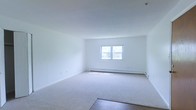This single-floor space presents a clean, minimalist layout designed for straightforward functionality. The main living area is finished with neutral wall-to-wall carpeting that transitions to hard flooring near the entryway, offering a practical combination of comfort and durability underfoot. Crisp white walls throughout the space create a bright, open atmosphere, while a centered window allows natural light to fill the room, enhancing the sense of openness. A baseboard heating system runs along the far wall beneath the window, providing an efficient and unobtrusive climate control solution. The smooth white ceiling complements the overall airy aesthetic, keeping the space feeling clean and uncluttered. The room's straightforward rectangular layout offers flexibility for a variety of furniture arrangements. With its neutral palette and well-maintained finishes, this space provides a blank canvas that is easy to personalize and adapt to different uses. The overall ambiance is calm and understated, making it well-suited for those who value simplicity and functionality in a living environment.