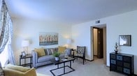 This single-floor residence offers a thoughtfully designed layout with a welcoming and functional living area at its heart. The living room is finished with plush wall-to-wall carpeting and painted in a soft, neutral tone that creates a bright and airy atmosphere. A textured ceiling adds subtle architectural character to the space. The room comfortably accommodates a contemporary gray sofa accented with warm yellow throw pillows, a sleek black geometric coffee table, and a patterned accent chair, together creating a cohesive and inviting seating arrangement. A framed cityscape artwork serves as a focal point on the main wall, adding a touch of urban sophistication. A stylish table lamp provides warm ambient lighting, complementing the overhead ventilation and lighting fixtures integrated into the ceiling. A doorway visible from the living area leads to additional rooms, hinting at a well-connected floor plan. The overall ambiance is calm, clean, and modern, with warm neutral tones and carefully chosen furnishings that make the space feel both livable and polished. The design strikes a balance between comfort and contemporary style, making it a versatile and welcoming environment.