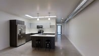 This single-floor space showcases a clean, industrial-modern aesthetic defined by polished concrete floors, crisp white walls, and an open layout that promotes a sense of spaciousness. The kitchen serves as the focal point, outfitted with stainless steel appliances including a full-size refrigerator, microwave, and range. Flat-front cabinetry in a muted gray tone lines the walls, complemented by light countertops that provide ample prep space. A dark-toned central island with a built-in sink and seating for two anchors the kitchen, adding both function and contrast to the overall palette. Pendant lighting hangs above the island, while recessed skylights and ceiling fixtures ensure the kitchen is well-illuminated. An exposed metal ductwork detail runs along the upper portion of one wall, nodding to the space's industrial character. Beyond the kitchen, an extended open area offers flexible use potential, continuing the polished concrete flooring and maintaining the minimalist aesthetic. A glass-paneled door at the far end allows natural light to filter into the space, enhancing the bright and airy atmosphere throughout.