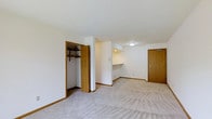 This single-floor space features a clean, functional layout with neutral white walls and warm wood-toned door and baseboard trim throughout. The flooring is covered in light-colored wall-to-wall carpet, lending a quiet, comfortable feel to the interior. A open closet with a built-in hanging rod and shelf provides practical storage, positioned conveniently off the main living area. A partial wall divides the space, creating a defined secondary area that includes a built-in desk or shelving nook—ideal for a workspace or study area. A second wood-toned door with a lever-style handle leads to an additional room or entry point, contributing to the overall flow of the unit. Ceiling-mounted flush lighting provides even illumination throughout. The overall design is straightforward and utilitarian, with an emphasis on functionality. The neutral color palette and consistent trim details create a cohesive look throughout the space, making it adaptable to a variety of uses and furnishing styles.