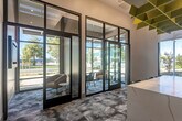 Located at 1240 La Avenida St in Mountain View, California, this contemporary commercial lobby delivers a polished and welcoming first impression through thoughtful architectural design and high-end finishes. The single-floor space is defined by soaring ceilings that amplify the sense of openness, while expansive floor-to-ceiling glass walls and black-framed curtain wall systems flood the interior with natural light and offer unobstructed views of the surrounding landscaped exterior.

A standout centerpiece is the elongated white marble-veined island, which serves as both a functional counter and a bold design statement. Overhead, a sculptural geometric ceiling installation in vibrant chartreuse yellow adds an artistic focal point and contributes to the space's acoustic performance. The patterned carpet flooring in soft blue-grey tones with an organic, fluid design grounds the space with texture and visual interest.

Along the perimeter, built-in cabinetry with a light wood finish provides practical storage, while a cushioned green banquette bench offers a comfortable seating area beside the glazed walls. Glass-enclosed alcoves near the entrance provide semi-private seating nooks with modern lounge chairs. The overall ambiance is bright, contemporary, and professional, making it well-suited for a leasing office, corporate reception, or community hub.
